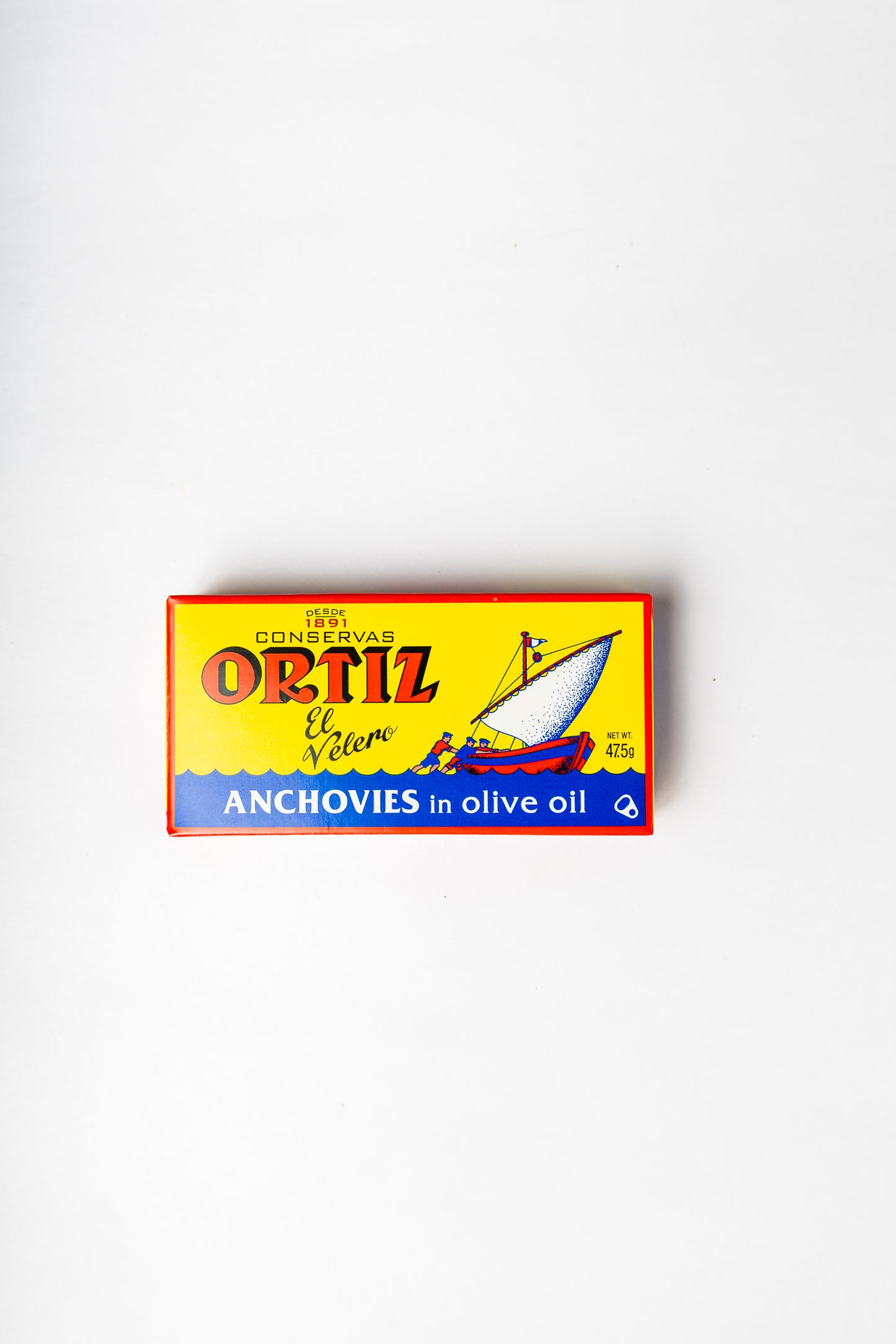 A rectangular box of Ortiz Confituras anchovies in olive oil, featuring a bright yellow background with a red border, blue band at the bottom, and an illustration of a sailboat, photographed on a plain white background.