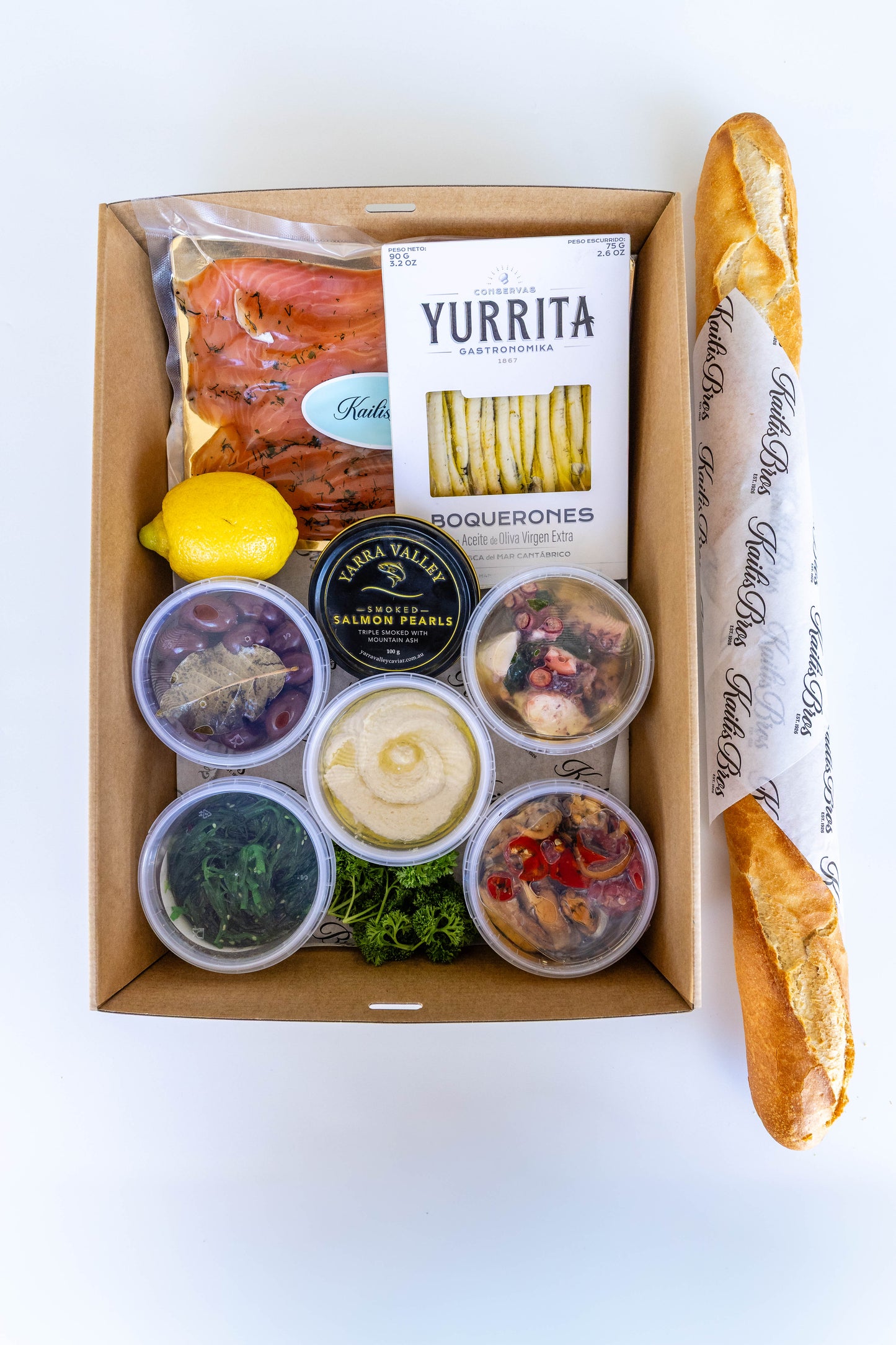 An overhead flat-lay of a gourmet food gift box on a white background, containing a baguette, Yurrita boquerones, salmon, hummus, olives, and various spreads, with a lemon and a small jar salmon caviar